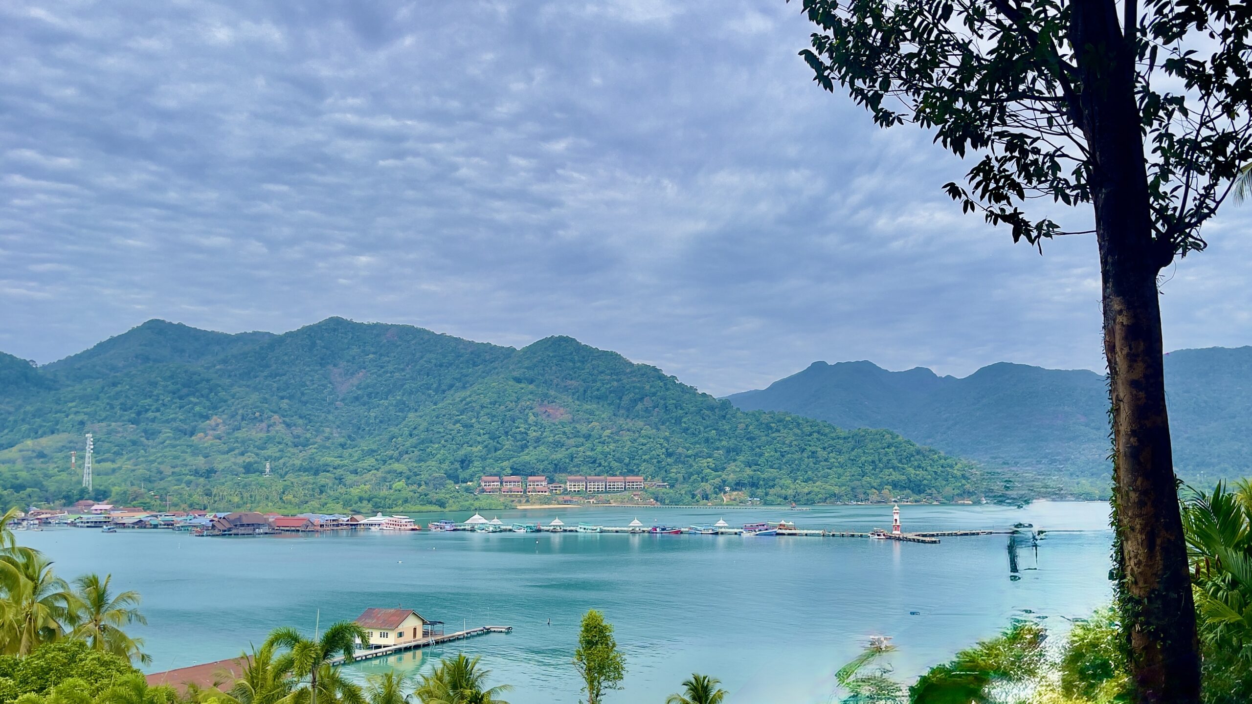 Scenic view of Bang Bao Bay in Koh Chang, Thailand, featuring turquoise waters, lush green mountains, and a charming fishing village with stilt houses and a white lighthouse. The tranquil bay, framed by tropical palm trees and a large tree in the foreground, highlights the island’s natural beauty and relaxed coastal atmosphere.