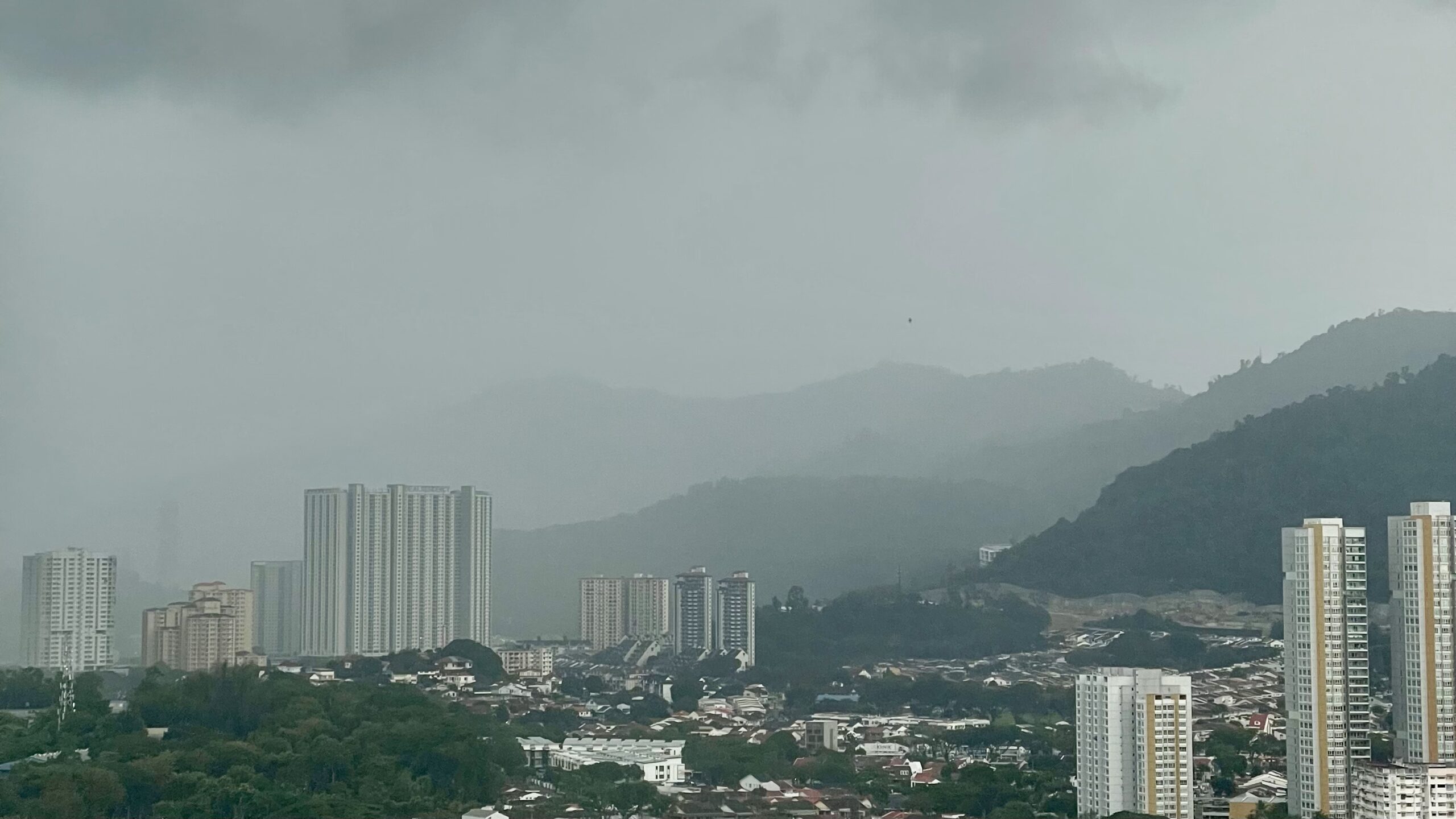 A rainy cityscape view from an apartment in Penang, Malaysia, featuring high-rise residential buildings, misty green hills, and an overcast sky. The rain clouds partially cover the mountainous landscape, creating a moody and serene atmosphere. This Penang city skyline captures the contrast between urban development and nature.