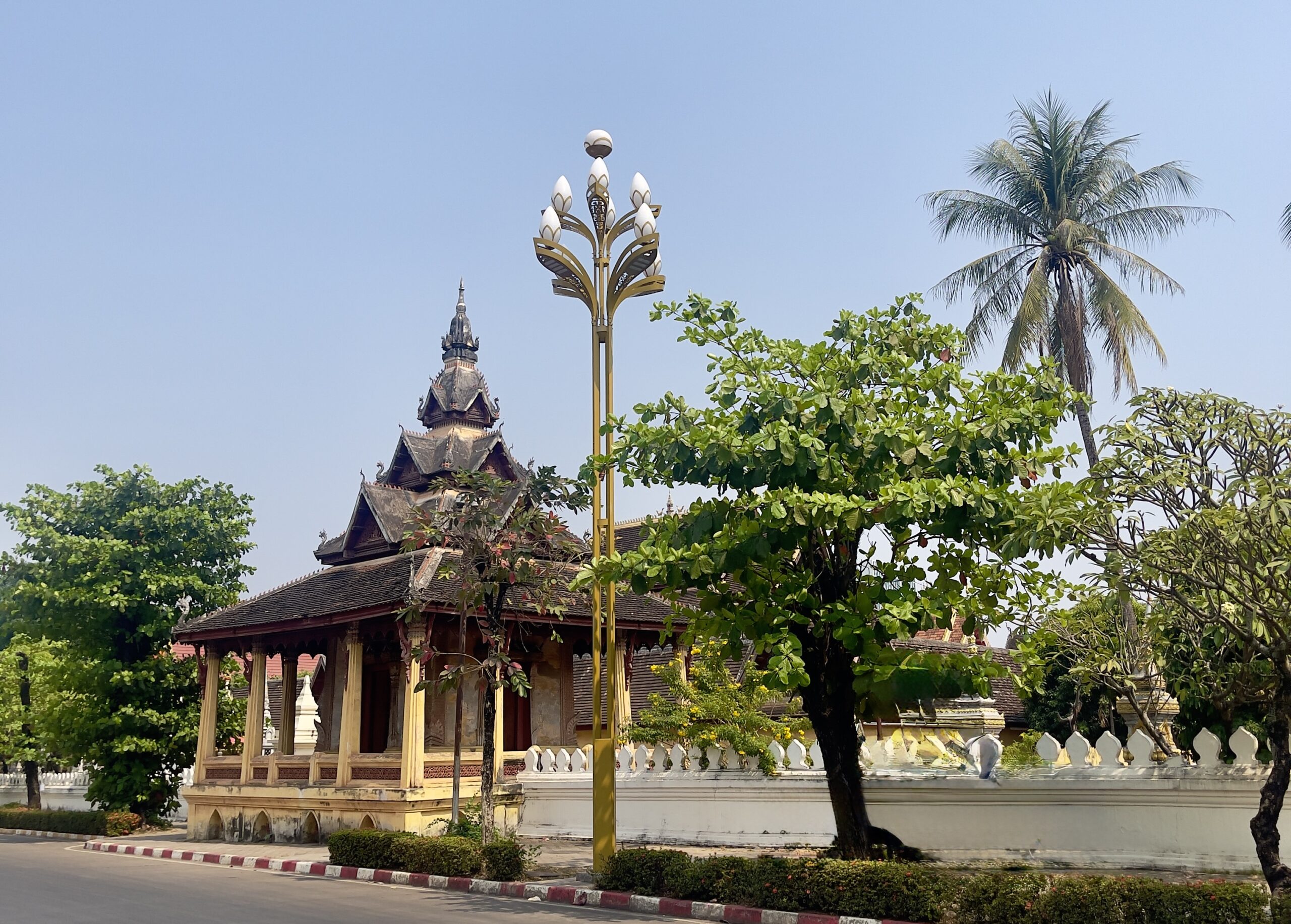 Traditional Lao temple with intricate wooden architecture in Vientiane, Laos, surrounded by lush greenery and a golden lotus-shaped street lamp.